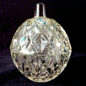 May include: A clear, faceted glass jar with a lid. The jar features a diamond pattern and a small, clear knob on top. It is likely used for storing small items or as a decorative piece.