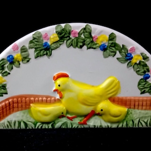 Può includere: Un elemento decorativo in ceramica a forma di semicerchio con una gallina gialla e due pulcini su un'area erbosa, con una staccionata marrone. Il bordo superiore presenta una ghirlanda floreale con foglie verdi, fiori blu, gialli e rosa.