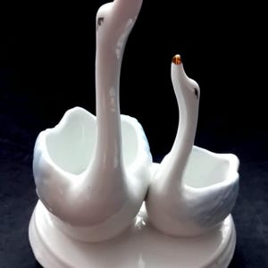 May include: A white ceramic swan-shaped egg holder with two compartments. The swans have long, curved necks and gold-coloured beaks. The holder is on a white, oval-shaped base. The background is black.