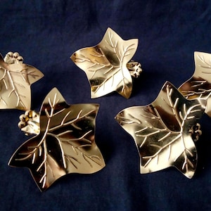 Op de afbeelding: Vijf goudkleurige, bladvormige decoratieve items. Elk blad heeft gedetailleerde nerven en een klein, getextureerd accent aan de basis. De items zijn gerangschikt op een donkerblauwe ondergrond, wat een opvallend contrast creëert.