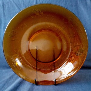 Op de afbeelding: Amberkleurige glazen plaat met reliëf bloemen- en sterpatronen. De plaat heeft een licht verhoogde rand en een glad, reflecterend oppervlak. De plaat wordt getoond op een zwarte metalen standaard, die het ontwerp en de kleur benadrukt.