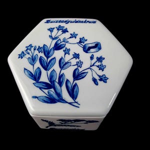 May include: A white, hexagonal ceramic trinket box with a blue floral design and the text "Suizonagoldenkruid". The box features a lid with a detailed illustration of blue flowers and leaves.