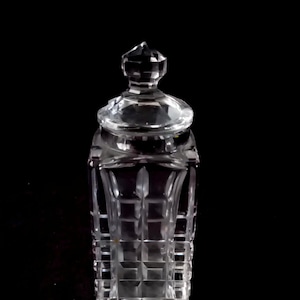 Può includere: Vaso di vetro trasparente e sfaccettato con coperchio. Il vaso ha una forma rettangolare con un motivo quadrato. Il coperchio è una versione più piccola della forma del vaso, con una parte superiore sfaccettata e appuntita. Il vaso viene probabilmente utilizzato per la conservazione o l'esposizione.