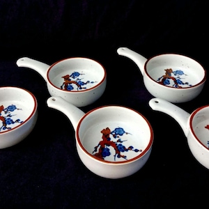 Può includere: Set di cinque piccole ciotole in ceramica bianca con manici. Ogni ciotola presenta un bordo rosso e un motivo floreale nei toni del blu e dell'arancione. Ideali per servire salse o piccole porzioni.
