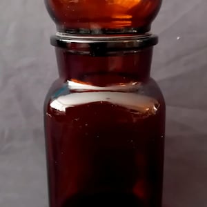 Op de afbeelding: Een donker amberkleurige apothekerspot van glas met een cilindrisch lichaam en een bijpassende ronde glazen stop. Het glas is doorschijnend en glad, met een brede hals en een dikke, gepolijste rand. Het ontwerp is eenvoudig en functioneel.