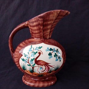 Op de afbeelding: Bruine keramische kan met een pauwenontwerp. De kan heeft een ronde body, een handvat en een tuit. De pauw is in rood, bruin en paars, met blauwe bloemen en groen blad. Een decoratief item.