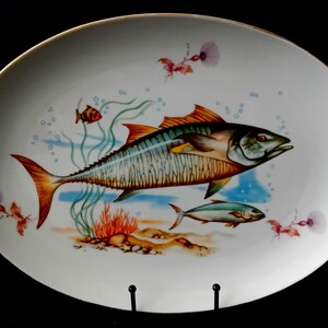 Może przedstawiać: Owalny biały półmisek ceramiczny z kolorową ilustracją dużej makreli, mniejszej ryby i maleńkiej pomarańczowej rybki pływających wśród zielonych wodorostów, pomarańczowego koralowca i jasnoniebieskich bąbelków na piaszczystym dnie morskim.