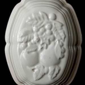 Op de afbeelding: Een ovale witte keramische wandvorm met een reliëf van twee peren omringd door bladeren en kleine bessen. De vorm heeft een gladde, glanzende afwerking en is voorzien van een klein gaatje aan de bovenkant om aan de muur te hangen.