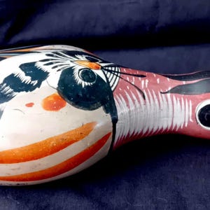 Puede incluir: Figurita de pájaro de cerámica pintada a mano. El pájaro tiene un pico rojo, ojos negros y blancos y un cuerpo con rayas naranjas y negras. Las alas están pintadas con detalles en blanco y negro. La figurita está sobre un fondo azul oscuro.
