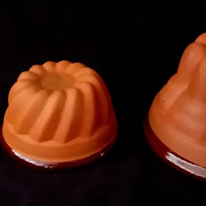 Op de afbeelding: Drie Bundt cakevormen in terracotta kleur van verschillende maten. Elke vorm heeft een gegroefd ontwerp en staat op een donker, reflecterend oppervlak. De vormen zijn gerangschikt van klein naar groot, waarbij hun verschillende capaciteiten worden getoond.