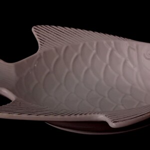 Pode incluir: Um prato de servir em cerâmica de cor clara em forma de peixe, apresentando um padrão detalhado de escamas em relevo no corpo e texturas lineares nas barbatanas e na cauda, apresentado contra um fundo preto sólido.