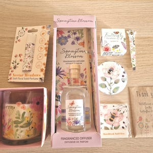 以下が含まれることがあります： 「Springtime Blossom」と「Nectar Meadows」の文字が入った、香りのディフューザー、キャンドル、香水、ネイルファイル、ワイルドフラワーの種など、花をテーマにしたアイテムのコレクション。 ピンク、紫、緑の色使い。