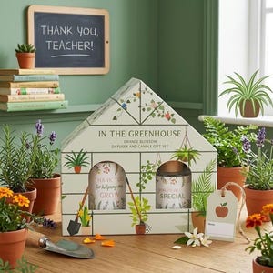 Puede incluir: Una caja de regalo con forma de invernadero etiquetada como IN THE GREENHOUSE ORANGE BLOSSOM DIFFUSER AND CANDLE GIFT SET. La caja presenta ilustraciones de plantas y herramientas, conteniendo dos recipientes con el texto THANK YOU for helping me GROW y you're so SPECIAL.