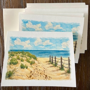 May include: Watercolor art prints of a beach scene with a sandy path, ocean, and blue sky with clouds. The prints feature a wooden fence and dune grass. The artist's signature is visible in the bottom left corner. Several blank envelopes are included.
