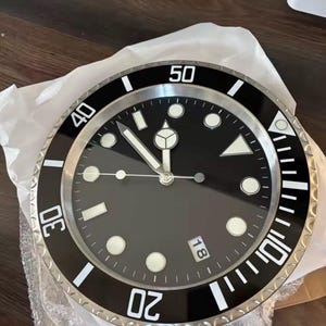 Peut inclure: Une horloge murale noire et argentée avec un cadran noir et des index blancs. L'horloge a une lunette argentée avec un anneau extérieur noir et des chiffres blancs. Les aiguilles de l'horloge sont argentées. La date est affichée à 3 heures.