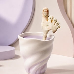 May include: A white ceramic cup with a swirling lavender design, filled with a lavender liquid, and a foot-shaped spoon. The spoon is off-white with detailed toes. The background features geometric shapes in shades of purple and pink.