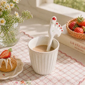 May include: A novelty ceramic spoon in the shape of a foot, with red nail polish, stirs a light brown drink in a white mug. A small cake with a strawberry and a basket of strawberries are also visible. The scene is set on a checkered tablecloth.