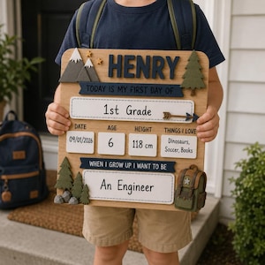 Könnte beinhalten: Ein hölzernes Schild für den ersten Schultag mit dem Namen Henry, dem Text 1st Grade sowie Feldern für Datum 09/01/2026, Alter 6, Größe 118 cm und Interessen wie Dinosaurier, Fußball und Bücher. Das Schild ist mit Berg- und Baummotiven verziert.