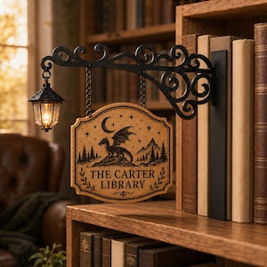 May include: A wooden sign hanging from a black metal bracket with an attached lantern. The sign features a black engraved dragon, mountains, trees, stars, and a crescent moon above the text THE CARTER LIBRARY, framed by decorative leaf motifs.