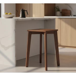 May include: A wooden stool with a square seat and four tapered legs. The stool is made of a dark-stained wood and has a simple, modern design. The stool is placed in a kitchen setting, near a countertop and cabinets.