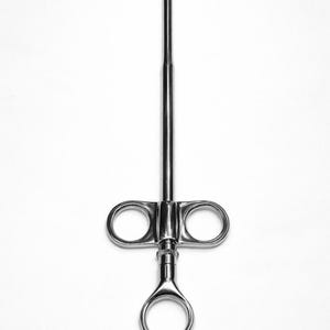 Pode incluir: Instrumento cirúrgico em aço inoxidável com um longo e fino eixo e uma pega com dois orifícios circulares para os dedos e um anel inferior. O instrumento é projetado para procedimentos médicos e está sobre um fundo branco.