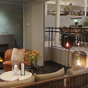 May include: A white ceramic mug with a black handle and the text "But First, Coffee" in a bold, black font. The mug is set against a blurred background, suggesting a cosy setting.