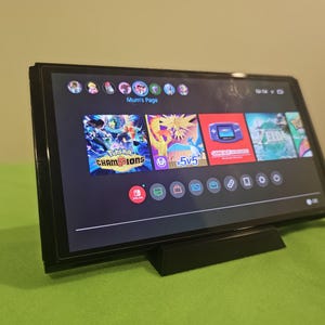 May include: A black Nintendo Switch console is displayed on a black stand, showcasing its home screen with game icons. The screen displays the text "Mum's Page" and various game titles, including "Pokemon Champions". The console is set against a green background.