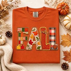 Op de afbeelding: Een roestoranje T-shirt met ronde hals en het woord FALL in grote, decoratieve letters. De letters zijn gevuld met verschillende patronen, waaronder patchwork, herfstbladeren, oranje gebreide texturen en een rood-zwart ruitpatroon.