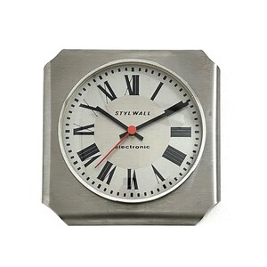 May include: A square wall clock with a brushed silver metal frame and a circular face. The clock features black Roman numerals, black hour and minute hands, and a thin red second hand. The text STYL WALL and electronic are printed in black.