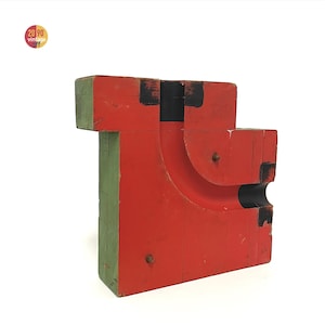 May include: A vintage, geometric wooden sculpture painted in red, black, and green. The blocky form features a curved cutout and is held together with metal hardware. The sculpture has a distressed, aged appearance, suggesting it is a vintage piece.