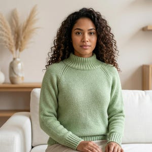 Może przedstawiać: Jasnozielony sweter z dzianiny z prążkowanym golfem i mankietami. Sweter ma rękawy raglanowe i wzorzysty wzór z dzianiny. Osoba siedzi na białej sofie.