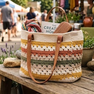 May include: A crocheted tote bag featuring horizontal stripes in cream, mustard yellow, burnt orange, and sage green. The bag has two tan leather handles attached with brass rivets and a small tag labeled HANDMADE. It contains a book and scarf.