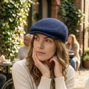 May include: A navy blue newsboy cap with a textured knit pattern. The cap has a rounded crown and a small, stiff brim. The woman in the image is wearing a cream-colored sweater. The background includes greenery and a brick building.