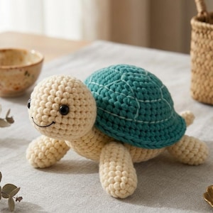 Puede incluir: Un juguete de tortuga de ganchillo con cabeza, patas y cuerpo de color crema, y caparazón turquesa. La tortuga tiene ojos negros y una boca sonriente. El caparazón tiene detalles de costura blancos. El juguete está sobre una superficie gris.