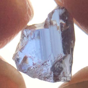 May include: A translucent, light purple gemstone with a raw, irregular shape and visible internal facets. The stone displays a pale violet hue and exhibits natural crystalline structures throughout its surface, catching light from behind.