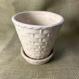 May include: A cream-colored ceramic plant pot with a raised basket-weave texture on the exterior. The pot features a matching circular saucer base and a wide, open top rim. The surface has a smooth, glazed finish with subtle variations in color.