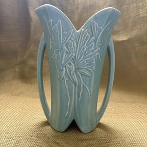 May include: A light blue ceramic vase featuring a raised relief design of two butterflies on the front. The vase has a tall, angular shape with two side handles and a V-shaped top opening. The surface has a smooth, matte finish in a solid color.