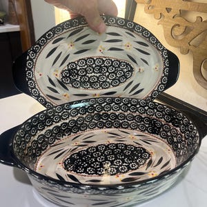 May include: Two oval ceramic baking dishes featuring a cream-colored base with black floral patterns, small yellow accents, and a dense black and white geometric border along the rim and center, with solid black handles on each side.