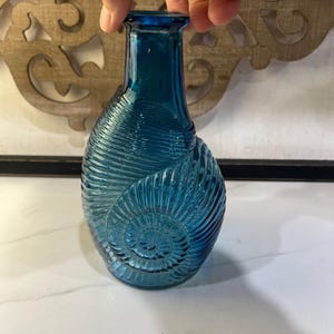 May include: A translucent deep blue glass vase featuring a prominent embossed spiral shell pattern that wraps around the body. The vase has a narrow neck and a flared rim, with a rounded base that tapers upward into a slender opening.