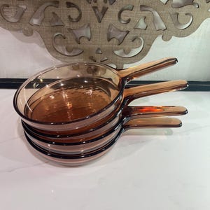 Op de afbeelding: Een stapel van vier ronde, amberkleurige glazen koekenpannen met geïntegreerde handgrepen. Elke pan heeft een gladde, transparante afwerking en een ondiepe, ronde kom, ontworpen met een consistente vorm en een uniforme bruine tint in het glas.