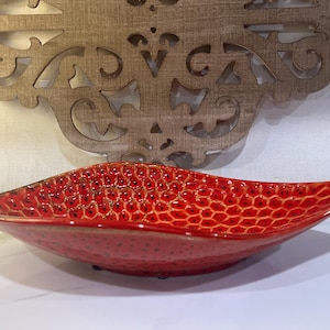 May include: A shallow, elongated ceramic bowl with a vibrant red glaze and a textured, honeycomb-like pattern across its interior surface. The bowl features dark, recessed spots within each hexagonal cell, creating a distinct contrast.