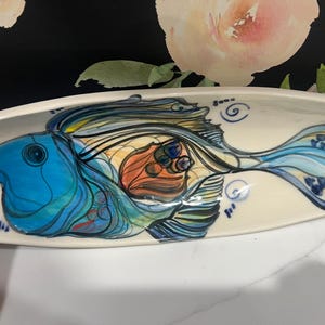 Puede incluir: Una fuente de cerámica ovalada con un diseño de pez pintado a mano. El pez tiene una cabeza azul, cuerpo naranja y cola azul y blanca. La fuente es de color crema.