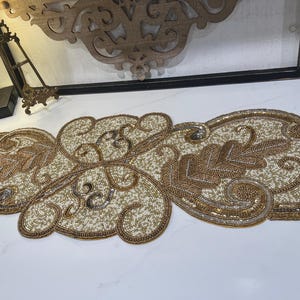 May include: A decorative table runner featuring intricate beadwork in shades of gold, silver, and cream. The design consists of swirling scroll patterns and leaf motifs, with a dense arrangement of small beads creating a textured surface.
