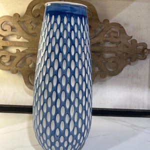 May include: A tall, cylindrical ceramic vase featuring a white base color with a repeating pattern of blue, elongated oval indentations. The top rim of the vase is finished with a solid blue band, creating a distinct contrast against the white interior.