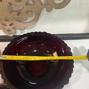 Könnte beinhalten: Eine tiefrote Glasschale mit gewelltem Rand und einem strukturierten Rautenmuster auf der Außenseite. Ein gelbes Maßband ist über die Oberseite gespannt und zeigt einen Durchmesser von etwa 24,8 cm für das runde Glasgefäß an.