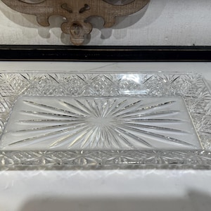 Könnte beinhalten: Ein rechteckiges Tablett aus klarem Glas mit einem zentralen Strahlenmuster, das von einem rautenförmigen Rand umgeben ist. Das Tablett hat ein facettiertes Kantendesign, das das Licht einfängt und eine reflektierende Oberfläche erzeugt.