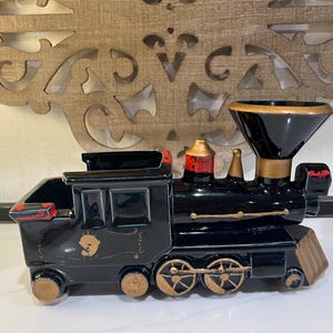 May include: A black ceramic train planter featuring a large funnel-shaped smokestack, gold-painted wheels, and red accents on the cab and boiler. The locomotive body has a hollow top section for planting and decorative gold line details on the side.