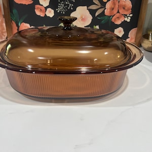 May include: Amber-coloured glass casserole dish with a matching lid. The dish has a ribbed exterior and a rounded shape. The lid has a knob handle. The dish is designed for baking and serving.