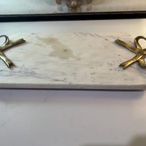 Op de afbeelding: Een rechthoekig marmeren serveerplank met goudkleurige strikhandvatten. De plank is wit met grijze adering. De handvatten zijn gevormd als strikken met hartvormige uitsparingen. Geschikt voor het serveren van hapjes of het presenteren van decoratieve items.