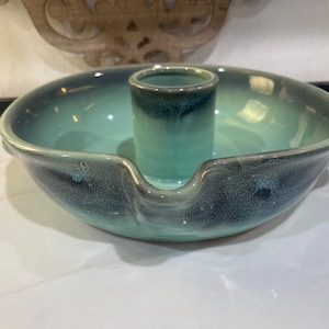 May include: A round ceramic baking dish with a central cylindrical pillar and a notched rim. The piece features a reactive glaze with a gradient of deep teal and dark blue tones, transitioning from a dark exterior to a lighter interior basin.